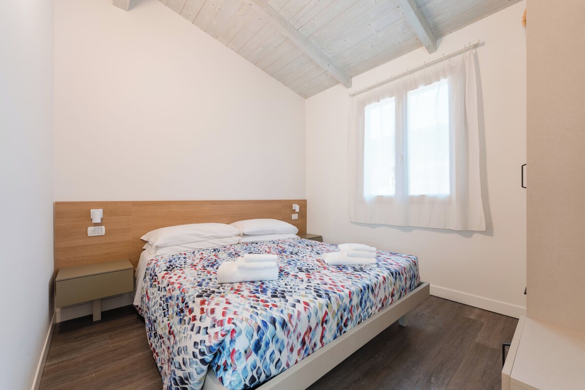 A single bed with a patterned bedspread, a bedside table, a nightstand, and a window with white curtains.