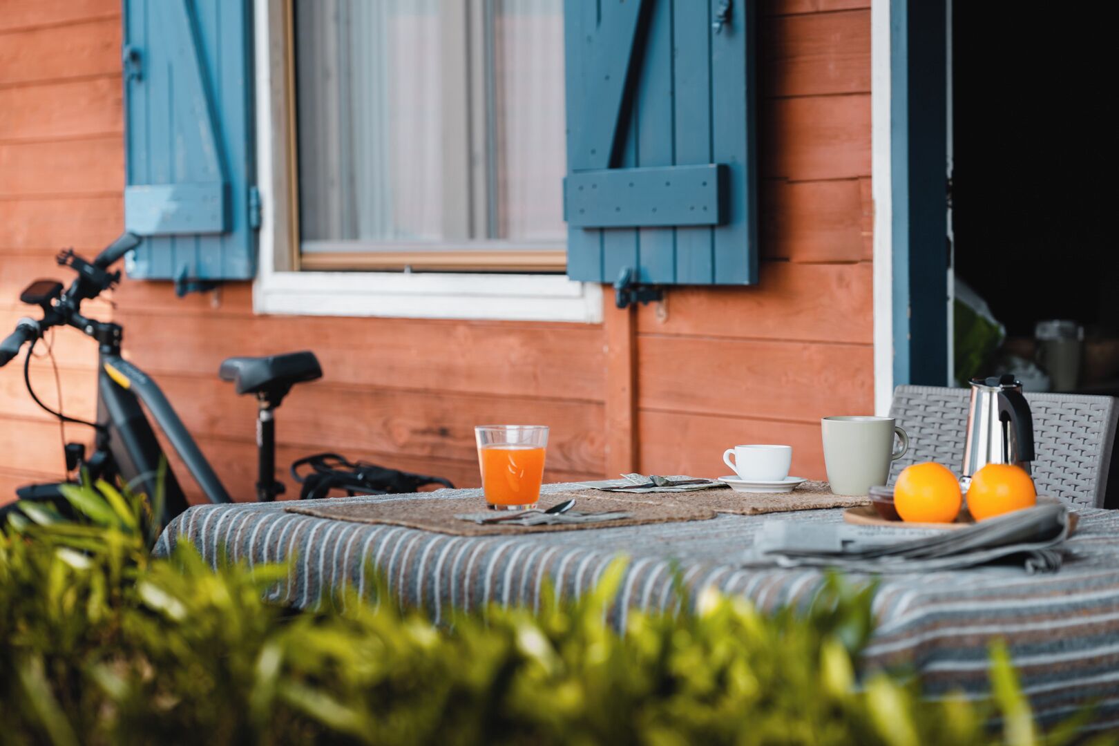 Ein Holzhaus mit blauen Fensterläden, ein Fahrrad, ein Glas Orangensaft, eine Tasse und zwei Orangen auf einem Tisch.