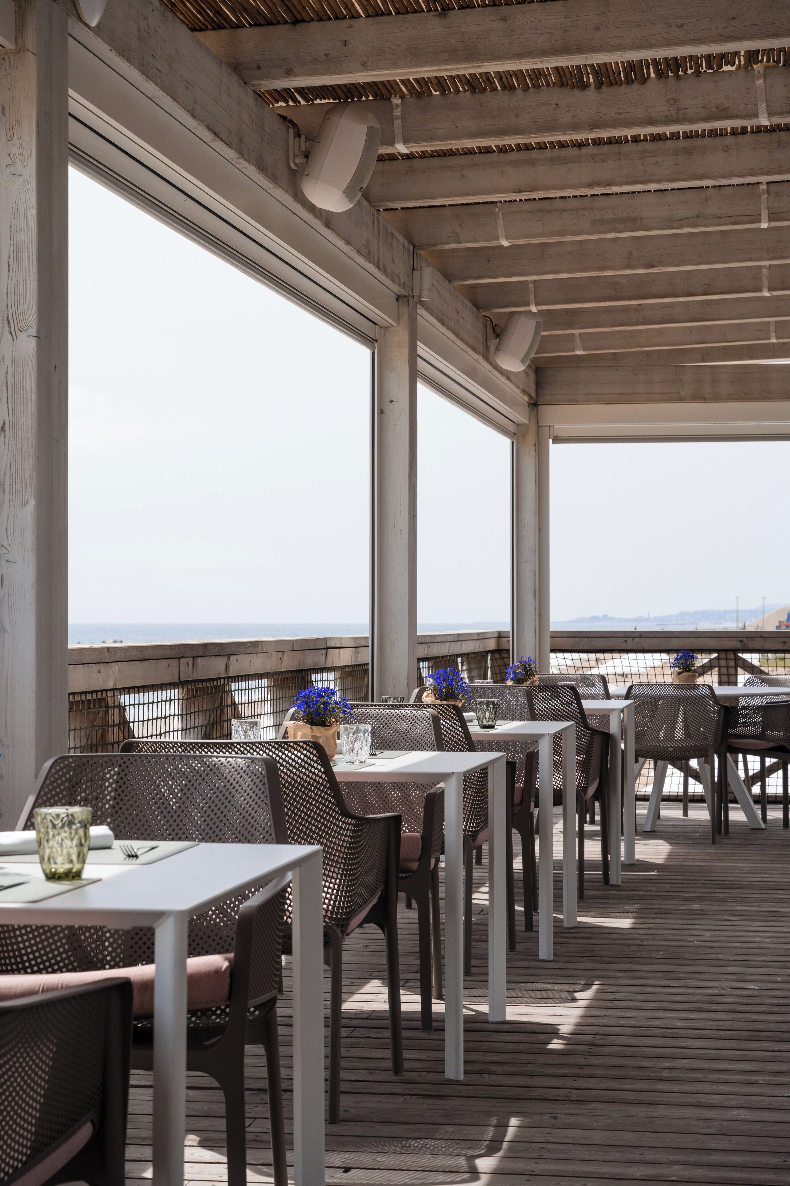 An outdoor dining area with tables and chairs, large windows, and a view of the ocean.