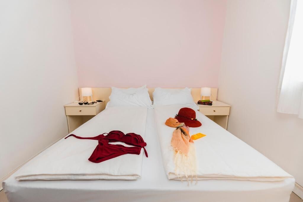 Un lit double avec du linge de lit blanc, deux tables de chevet avec des lampes et un vêtement rouge sur le côté gauche.