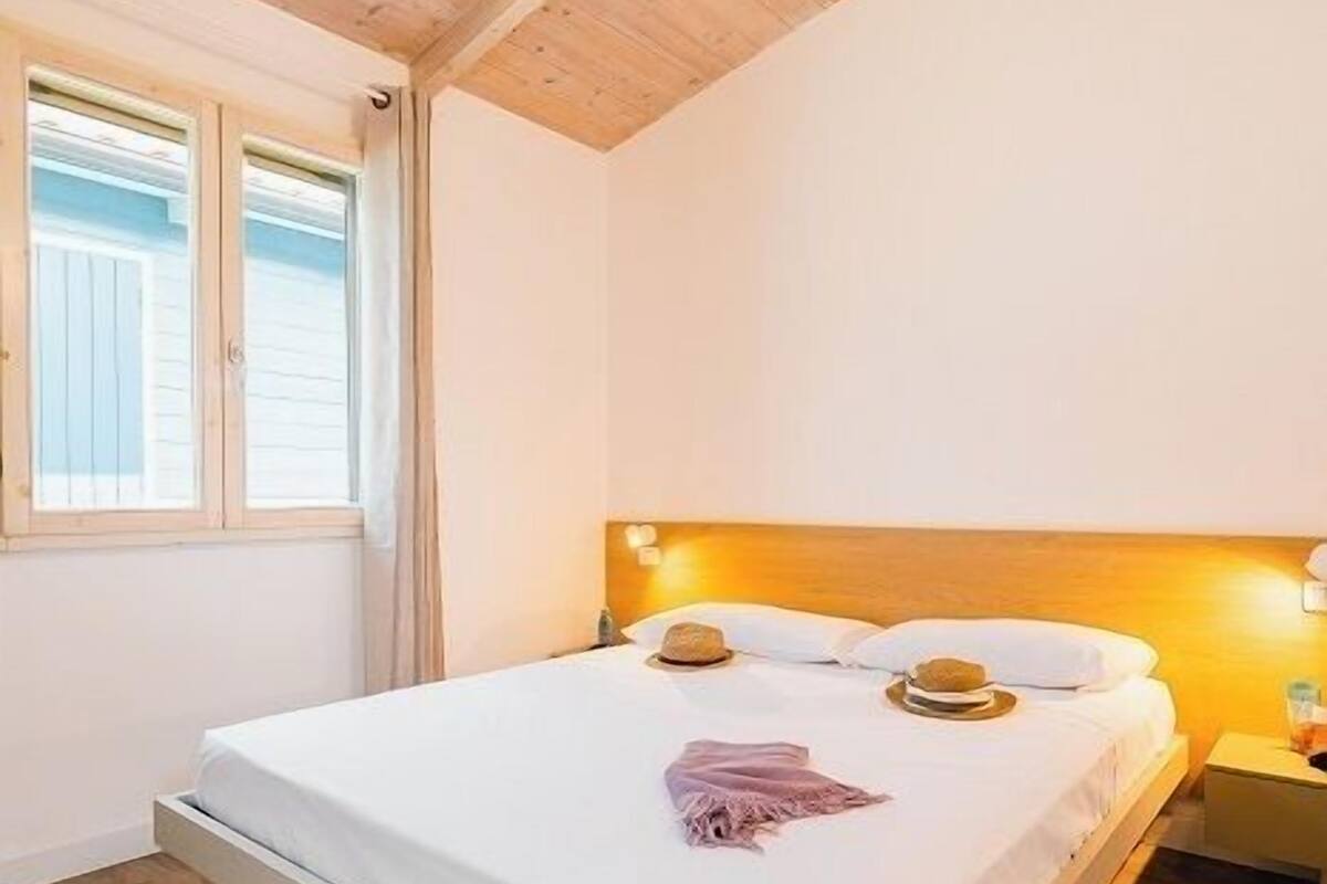 A bedroom with a wooden ceiling, a bed with white bedding, a bedside table with a lamp, and a window with curtains.