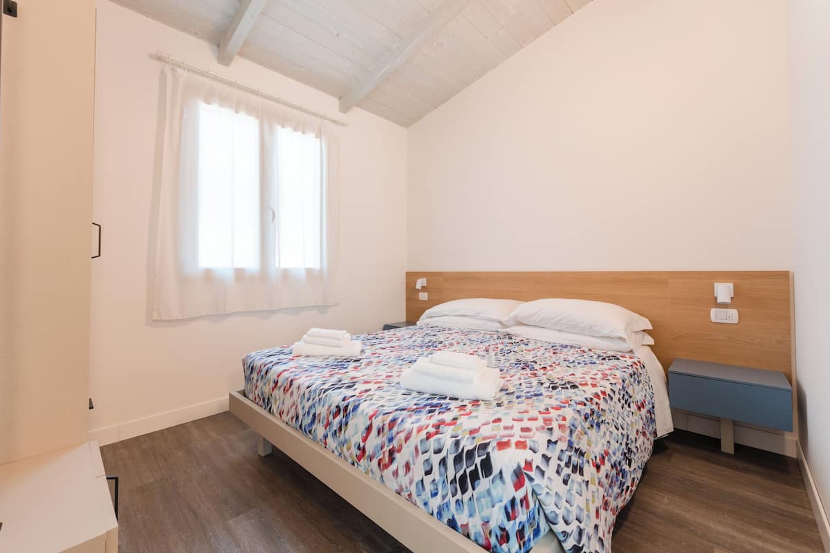 A single bed with a patterned bedspread, a bedside table, a nightstand, a window with curtains, and a wooden ceiling.