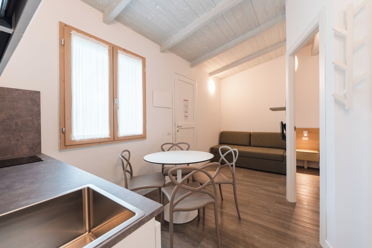 A modern kitchen with a sink, dining table, and chairs. There is a sofa and a lamp in the living area.
