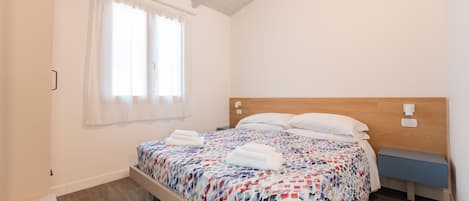 Habitación individual con una cama que tiene cabecera de madera, una mesita de noche y una ventana con cortinas blancas.