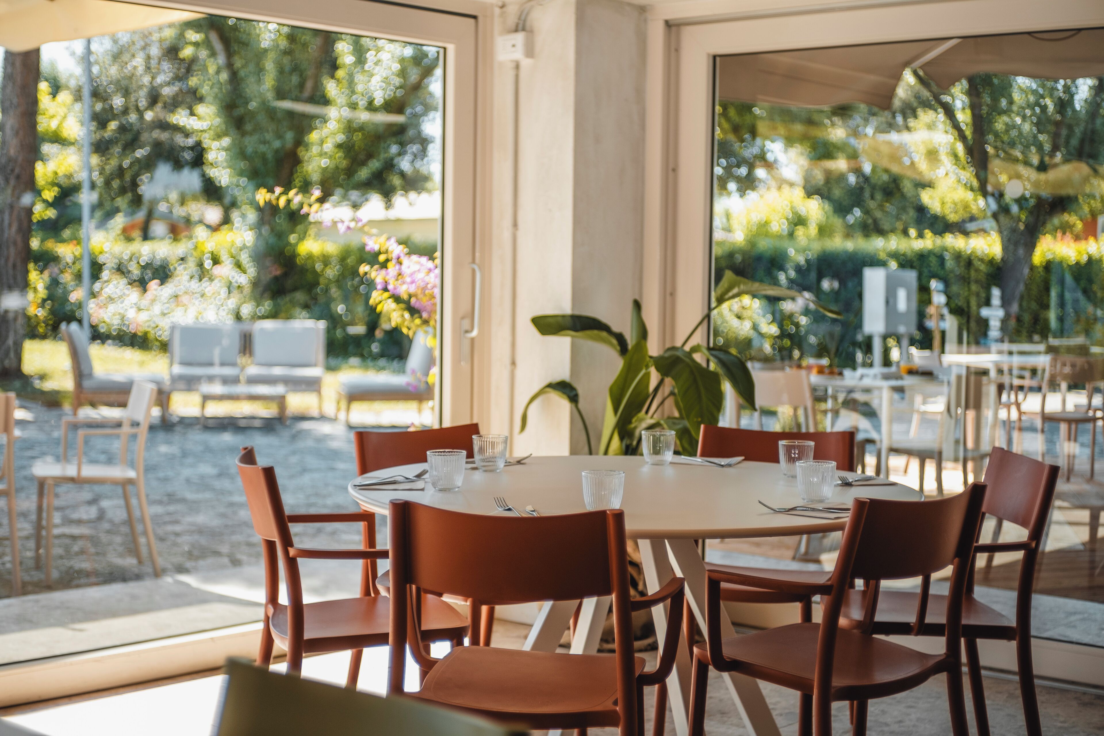 An outdoor dining area with a table set for two, surrounded by chairs, with a view of greenery and flowers through the glass.