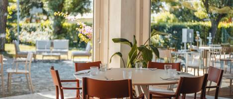 An outdoor dining area with a table set for two, surrounded by chairs, with a view of greenery and flowers through the glass.