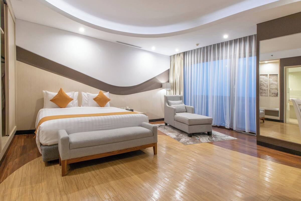 Una habitación de hotel moderna con una cama grande, un sofá, una silla y un amplio ventanal con cortinas.