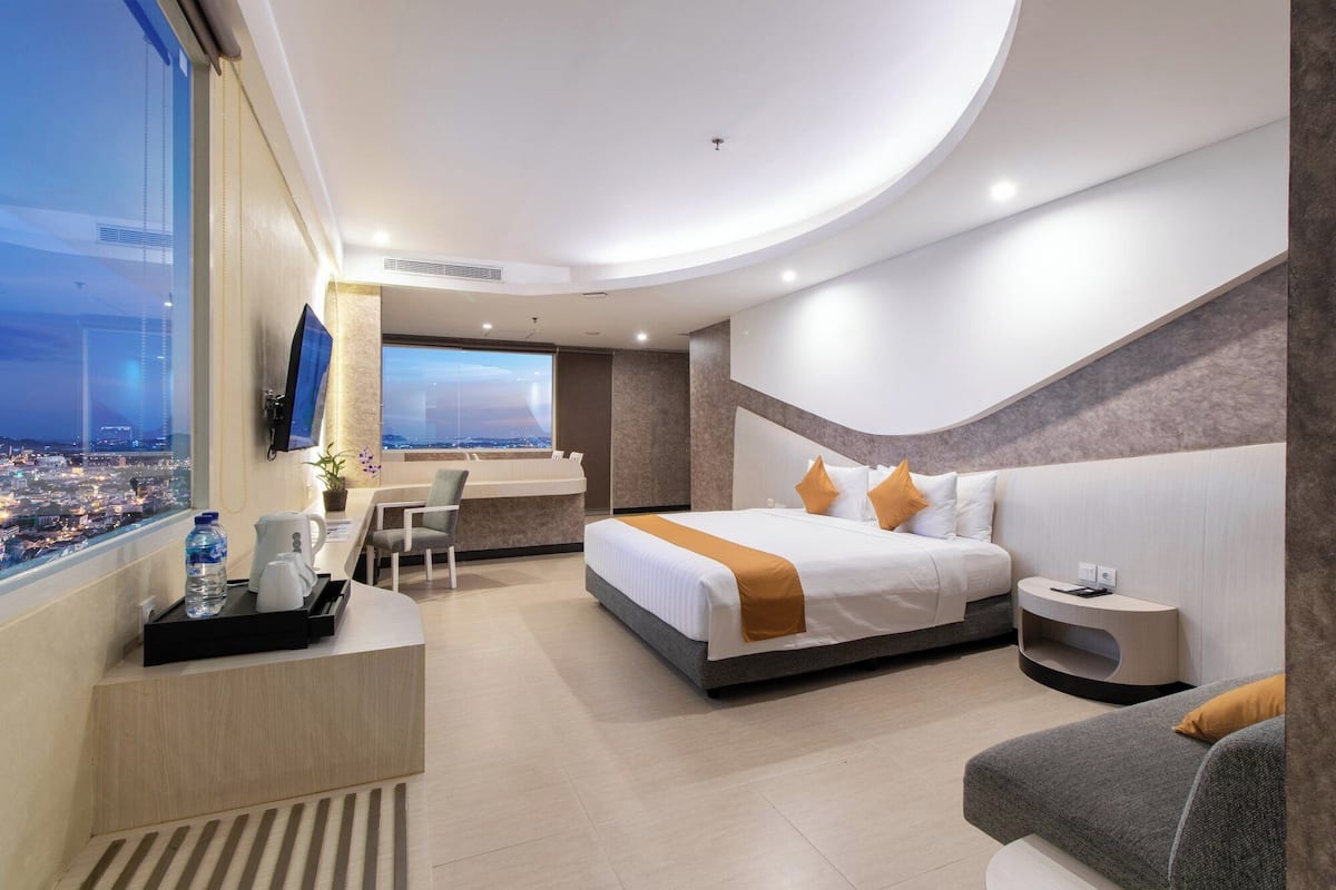 Una habitación de hotel moderna con una cama grande, un escritorio, una silla y vistas a un paisaje urbano a través de un ventanal.