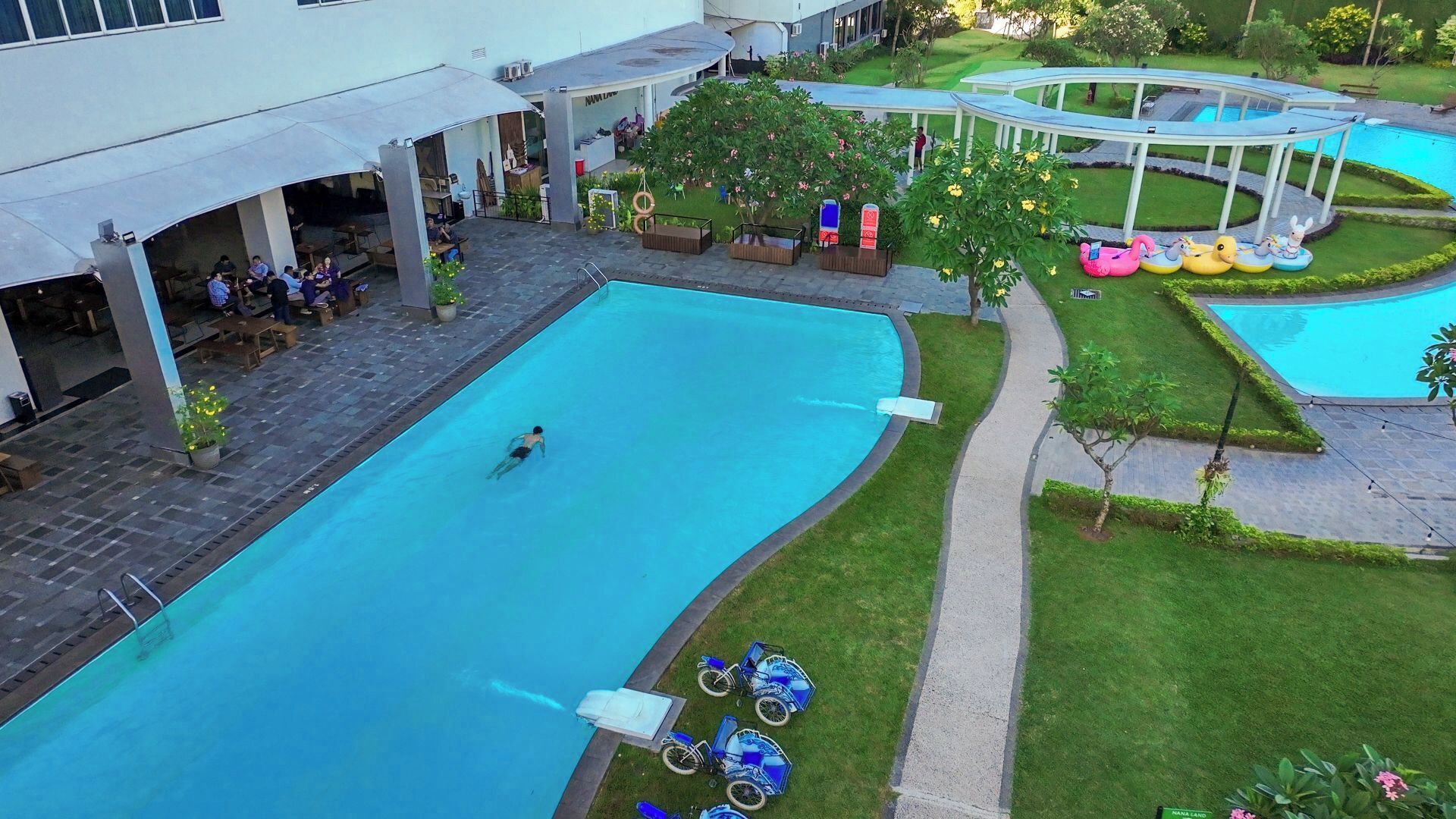 A swimming pool area with a person swimming, lounge chairs, and a covered seating area.