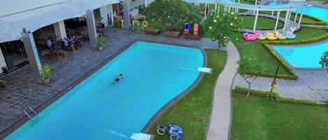 A swimming pool area with a person swimming, lounge chairs, and a covered seating area.