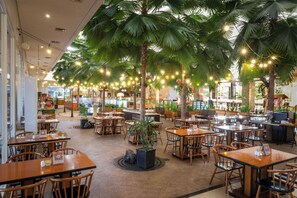 Un restaurante con mesas y sillas de madera, rodeado de palmeras y luces colgantes.