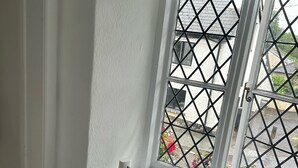 Una habitación con ventana que tiene una celosía metálica, una pared blanca y un pequeño florero blanco en el alféizar.