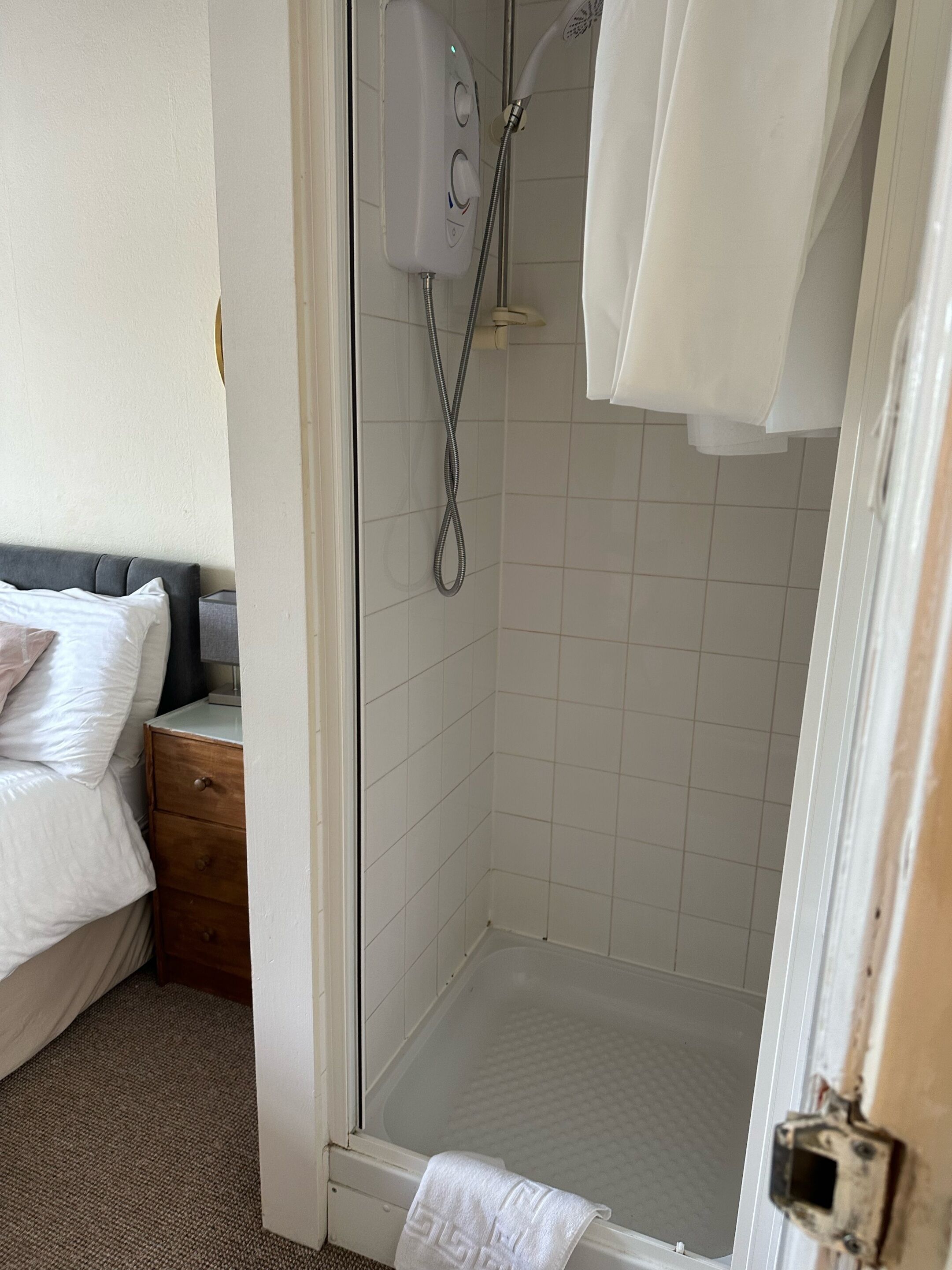 A hotel bathroom with a glass-enclosed shower, a white bathtub, and a wooden nightstand with a drawer.