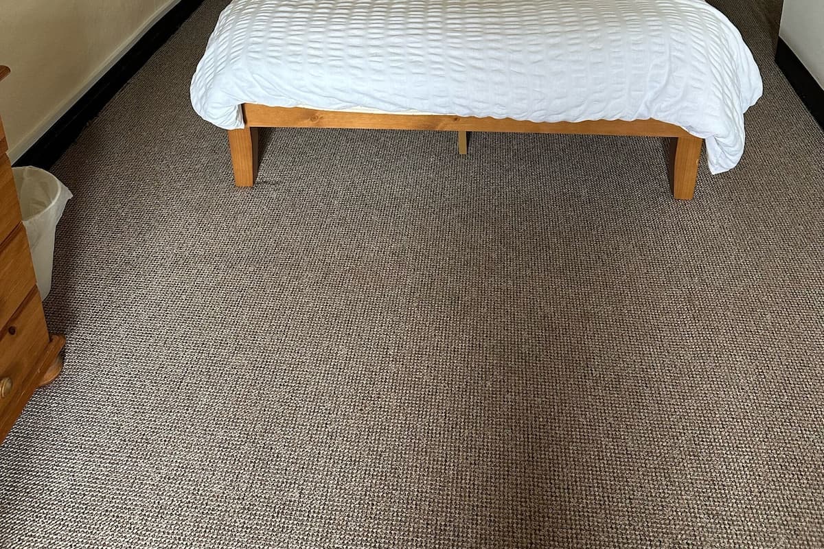 A neatly made bed with a white comforter and two pillows, one of which is orange. There are two bedside tables with lamps, and a wooden nightstand with a lamp. The room has a carpeted floor and a plain wall with a small window.