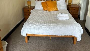 Una cama bien hecha con una colcha blanca y dos almohadas, una de ellas naranja. Hay dos mesitas de noche con lámparas, y un cabecero de madera con una lámpara. La habitación tiene piso alfombrado y una pared lisa con un ventanal pequeño.