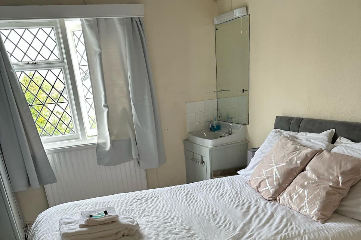 A neatly made bed with a white comforter and two pillows, a small bedside table with a mirror, a window with curtains, and a radiator.