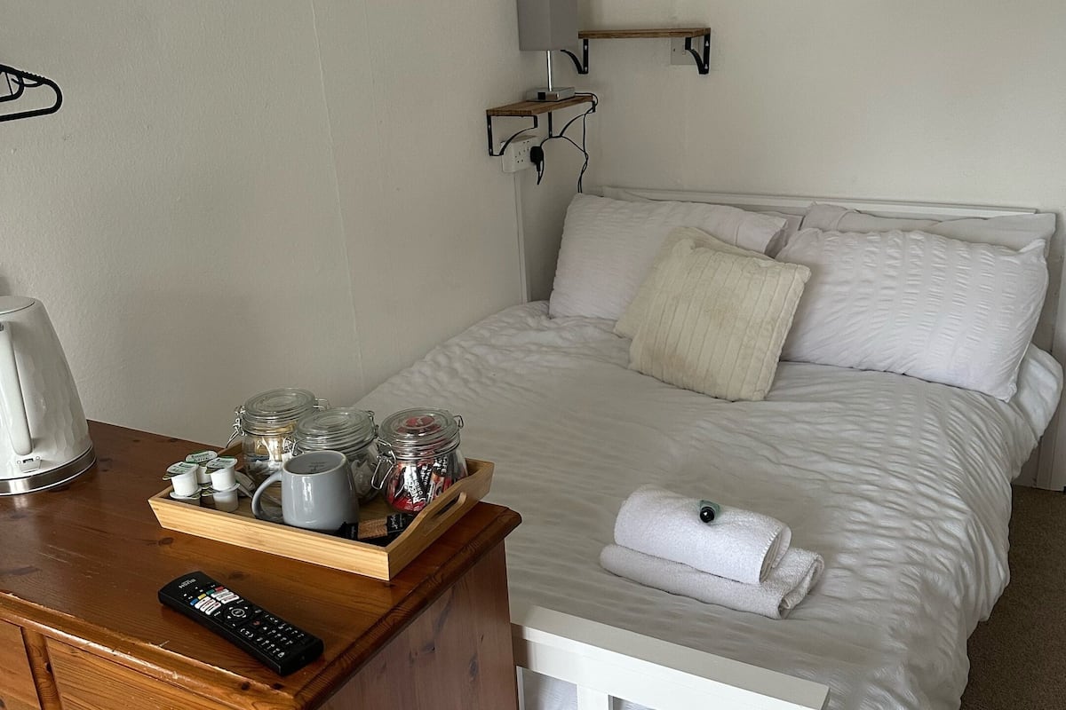 A neatly made bed with white linens, a bedside table with a remote control, a tray with jars and mugs, and a wall-mounted lamp.