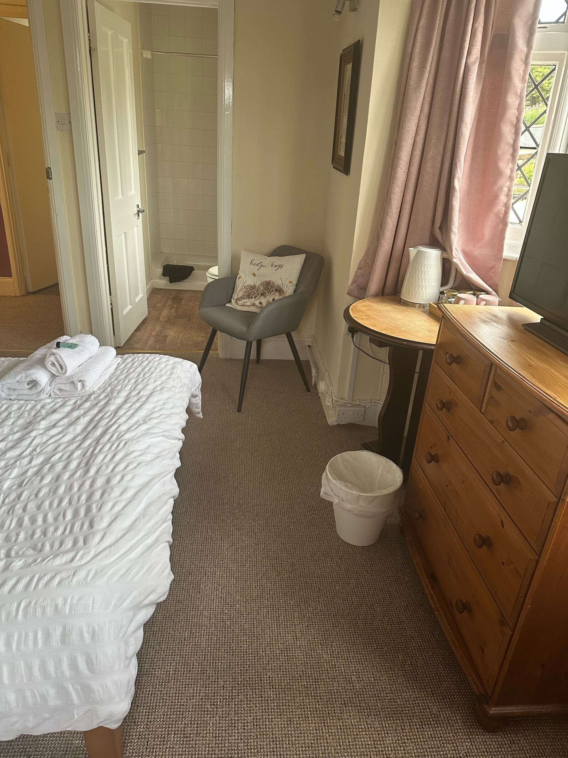 A bedroom with a bed, a chair, a dresser, a bedside table, a window with curtains, and a door leading to a bathroom.