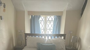 Una cama bien hecha con edredón blanco y dos almohadas, una ventana con cortinas azules y dos lámparas de noche.