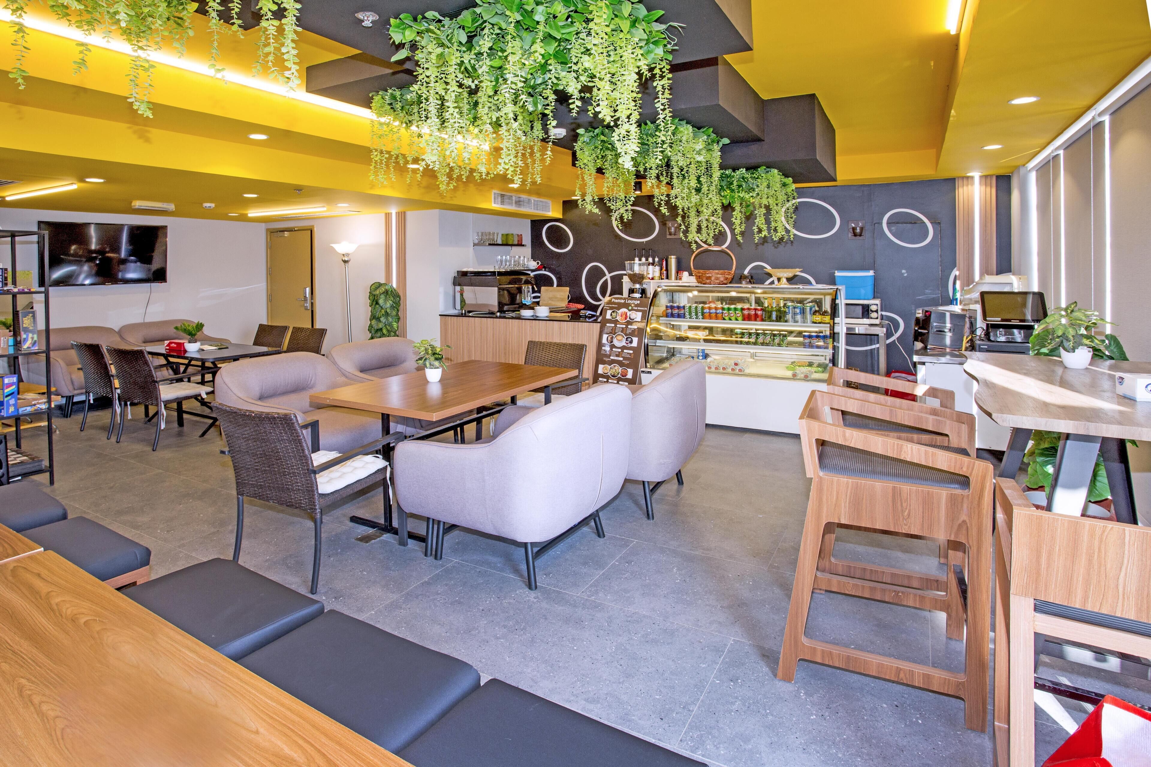 A modern restaurant with a central dining area featuring a wooden table and cushioned seating, surrounded by a kitchen area with a coffee machine and a refrigerator.
