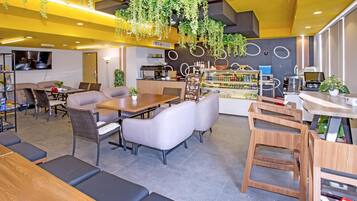 A modern restaurant with a central dining area featuring a wooden table and cushioned seating, surrounded by a kitchen area with a coffee machine and a refrigerator.