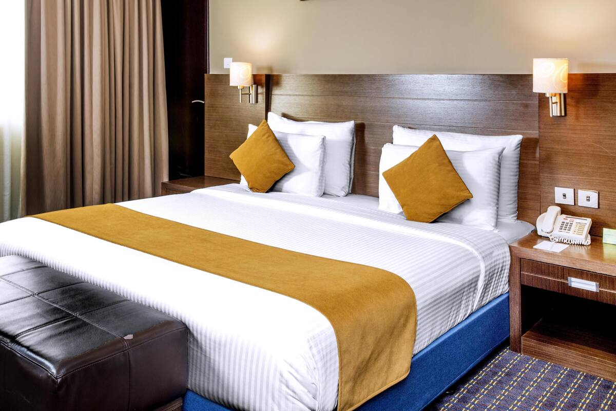 A hotel room with a large bed, a wooden headboard, two bedside lamps, a bedside phone, and a brown leather ottoman.