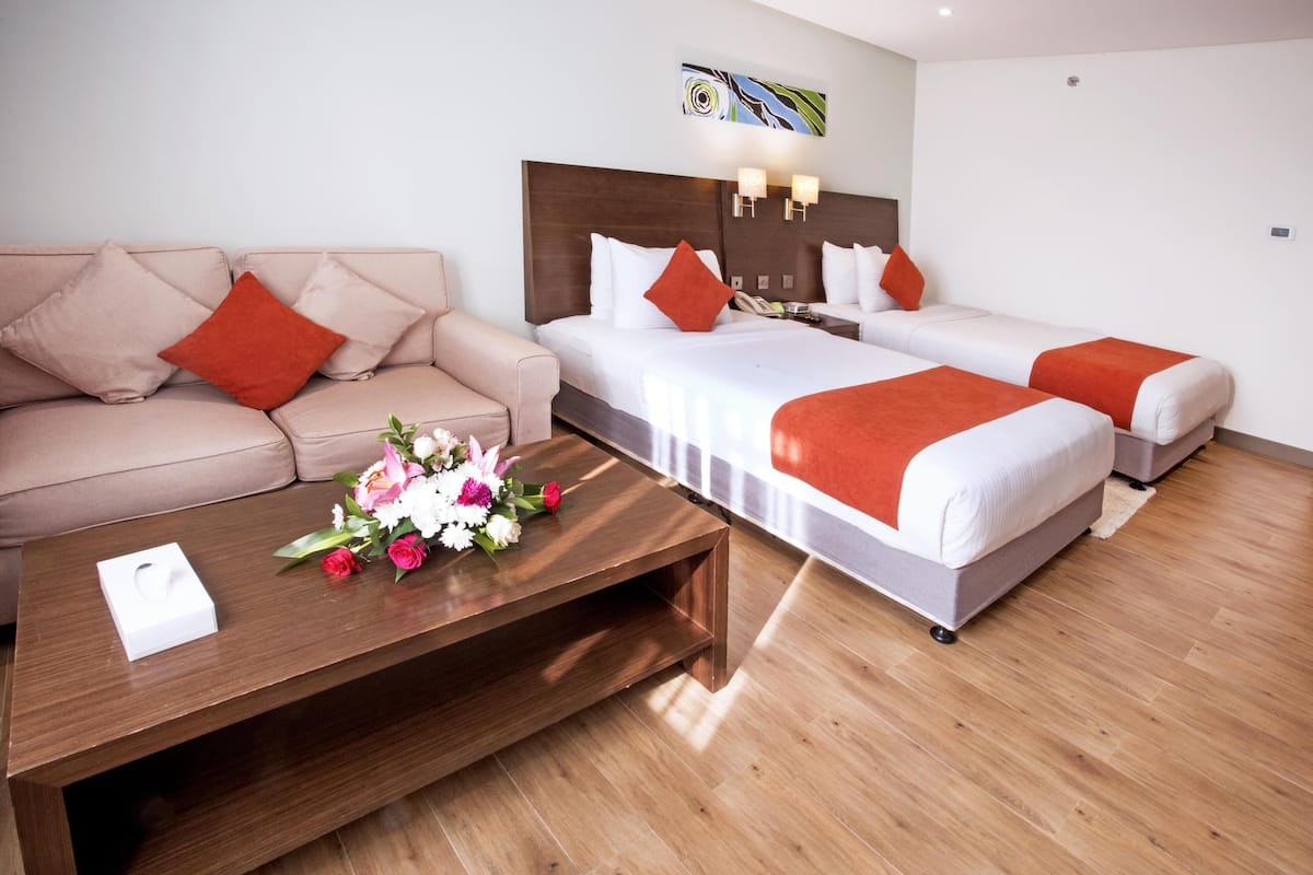 A hotel room with a sofa, two beds, a wooden coffee table with flowers, and a wall-mounted light fixture.