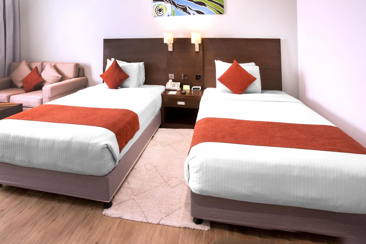 A hotel room with two beds, a sofa, a nightstand with a phone, and a wall art piece.