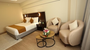 Habitación de hotel con cama, dos sillones, una pequeña mesa de madera con un florero y una mesita de noche con un teléfono.
