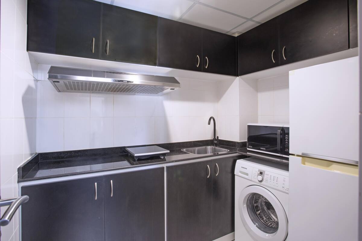 A compact kitchen with dark cabinets, a stainless steel range hood, a microwave, and a built-in washing machine.