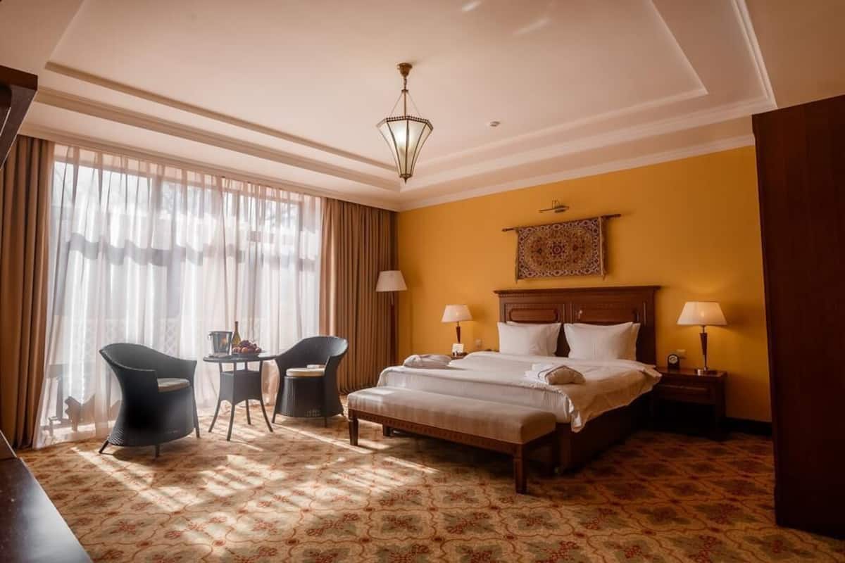 A hotel room with a large bed, two bedside tables with lamps, a small table with chairs, and a large window with curtains.