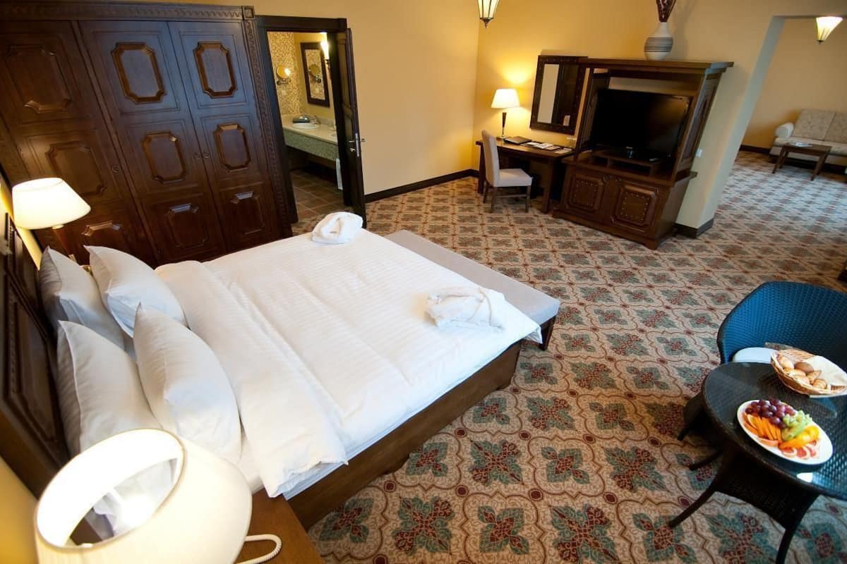 A hotel room with a large bed, a desk with a television, a small table with fruit, and a patterned carpet.