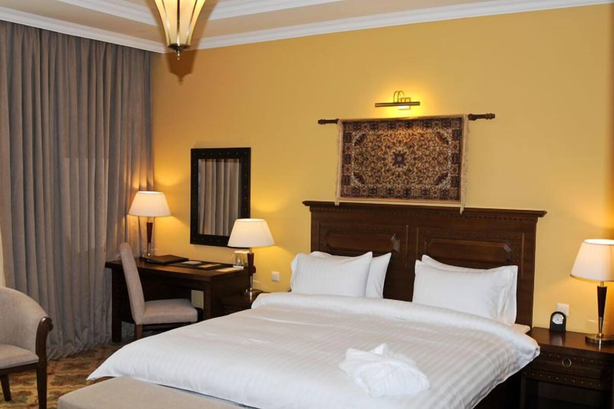 A hotel room with a large bed, two bedside tables with lamps, a desk with a chair, and a patterned tapestry above the bed.
