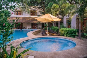 Une piscine entourée de chaises longues et d’un parasol, le tout bordé d’une végétation luxuriante et adossé à un bâtiment dont le toit est fait de chaume.