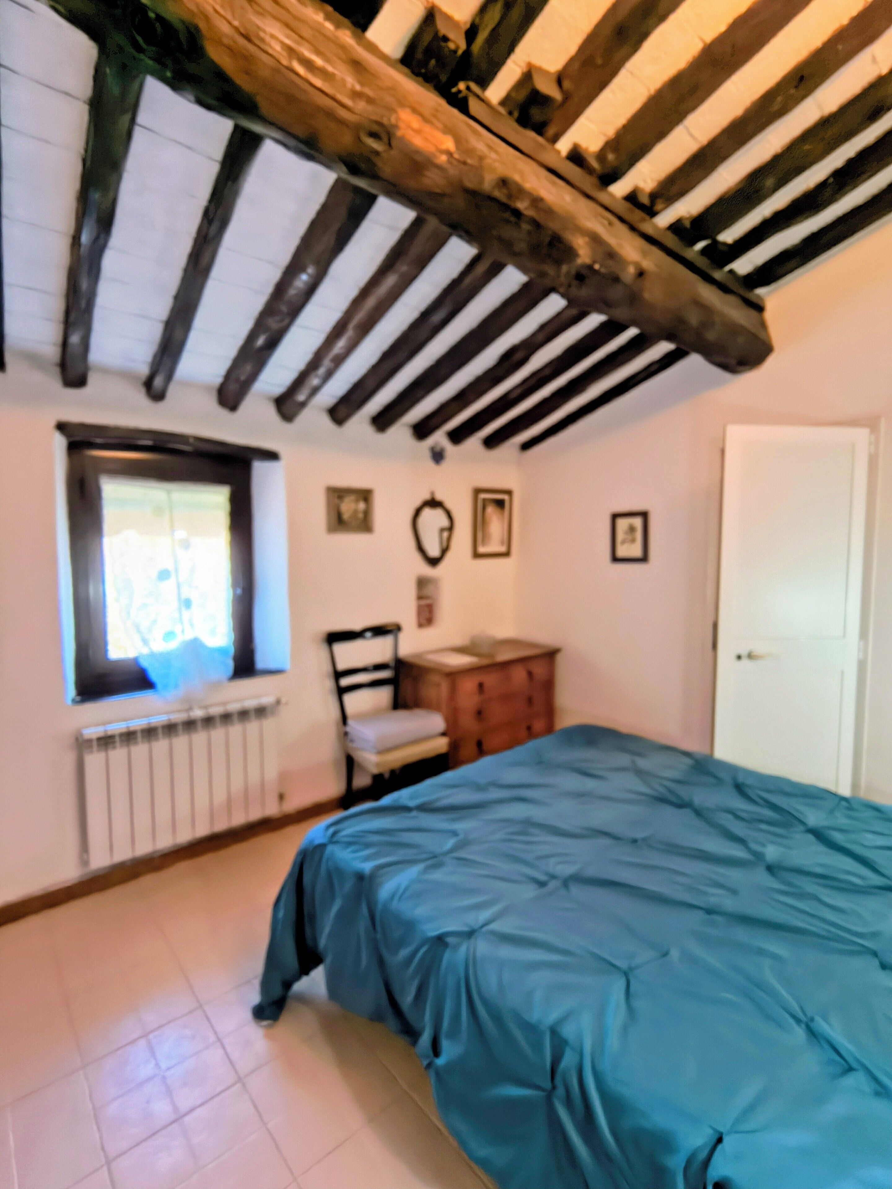 A bedroom with a bed, a window, a radiator, a wooden chair, a dresser, and a door.