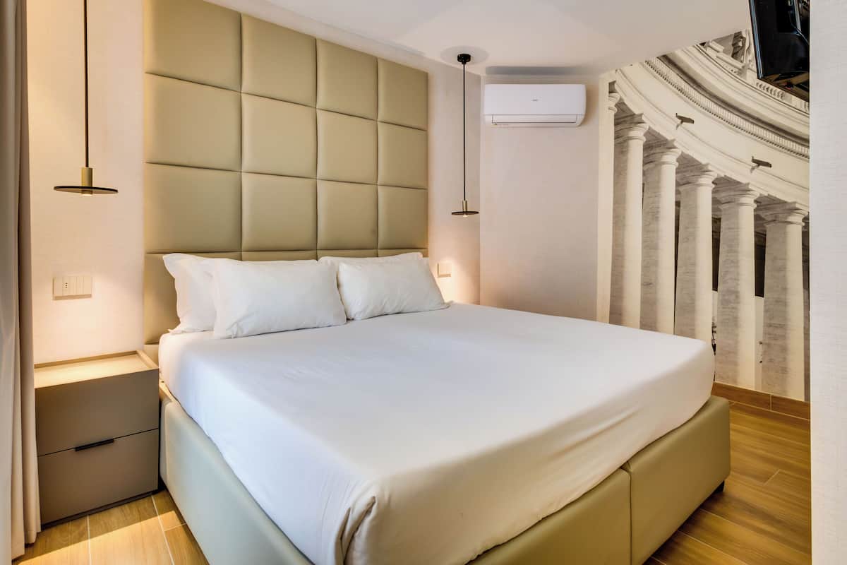 A modern hotel room with a large bed, a headboard with a tufted design, bedside lamps, and a wall-mounted air conditioning unit.