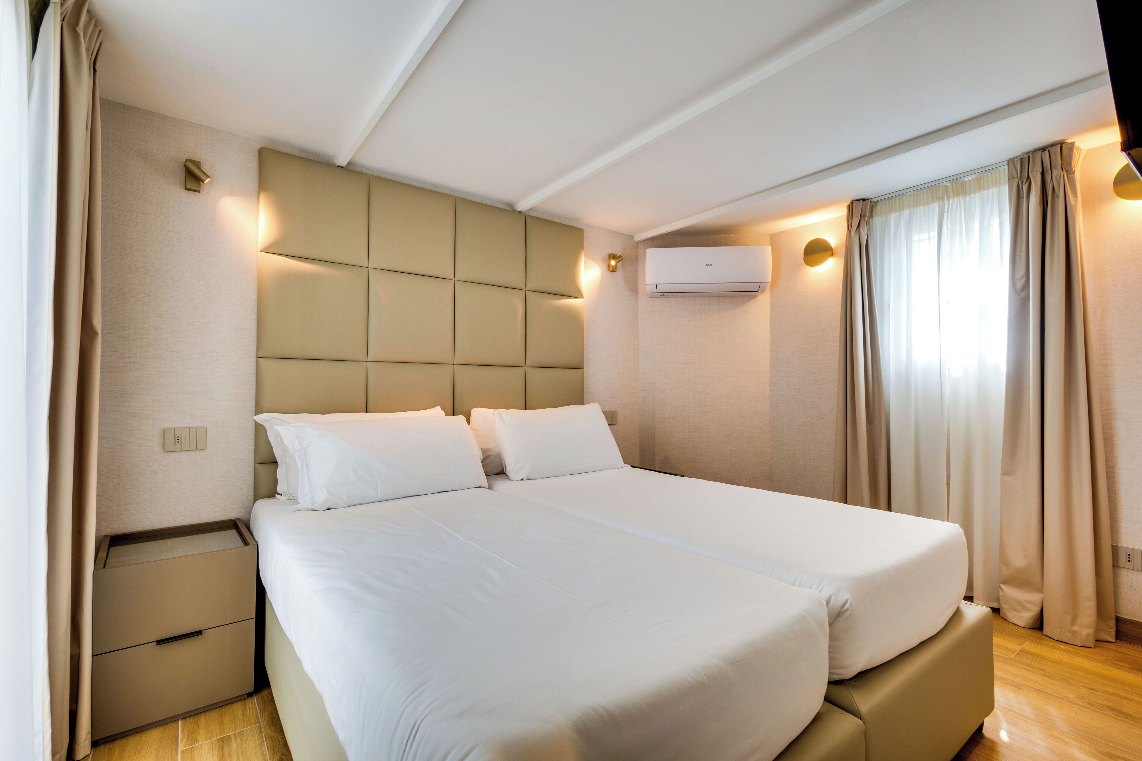 A modern hotel room with a large bed, a headboard with illuminated panels, a bedside table, and an air conditioning unit.