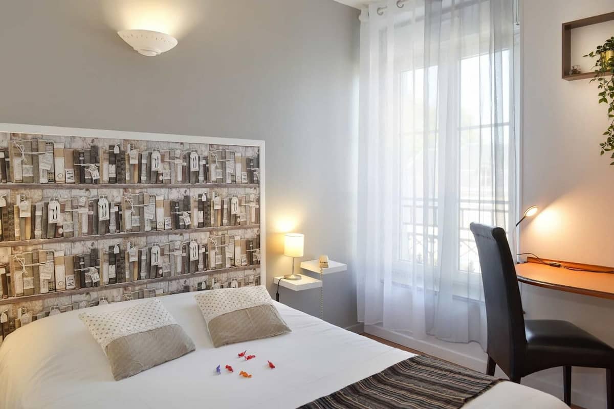 A hotel room with a large bed, a desk with a chair, a wall-mounted shelf with various items, and a window with sheer curtains.