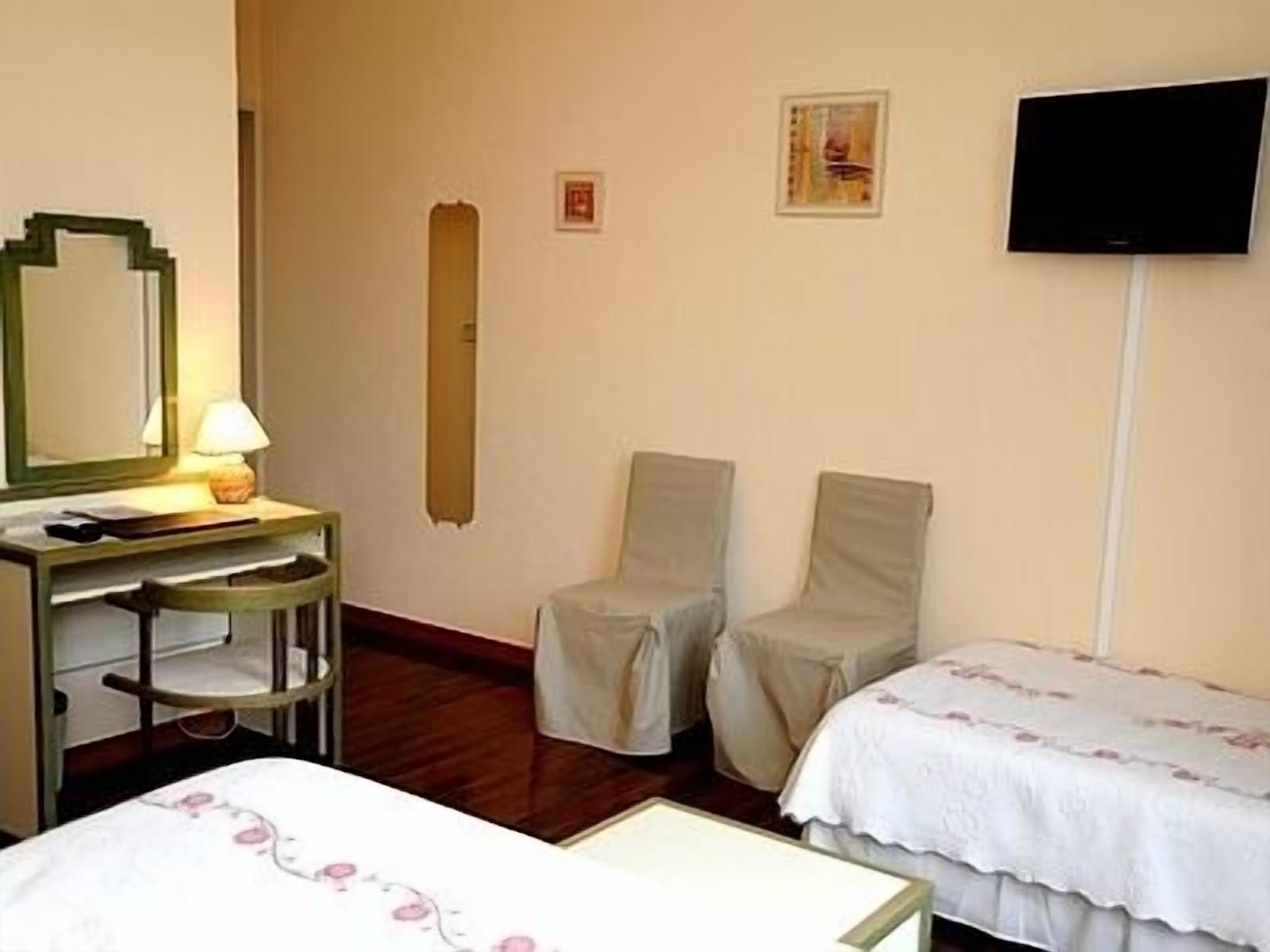 A hotel room with a bed, a desk with a lamp, two chairs, a television on the wall, and a small painting.