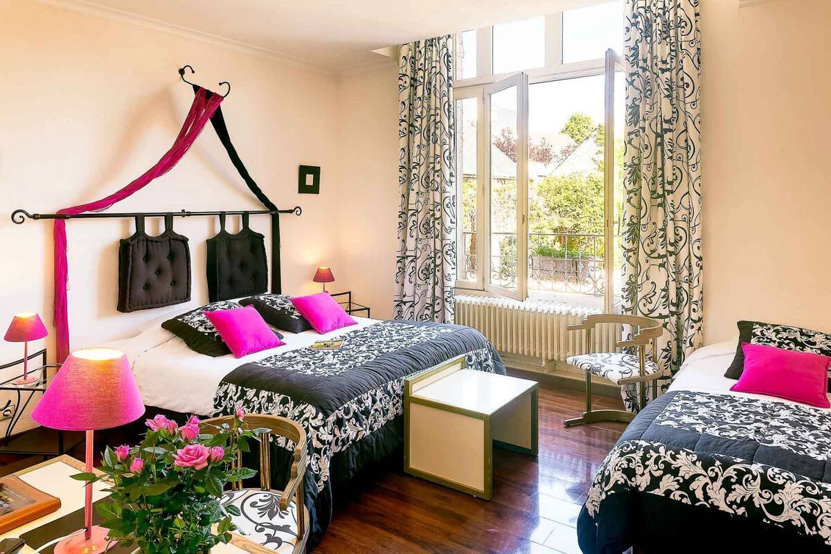A bedroom with a four-poster bed, two bedside tables, a small bedside lamp, a chair, and a view of the garden through a window.