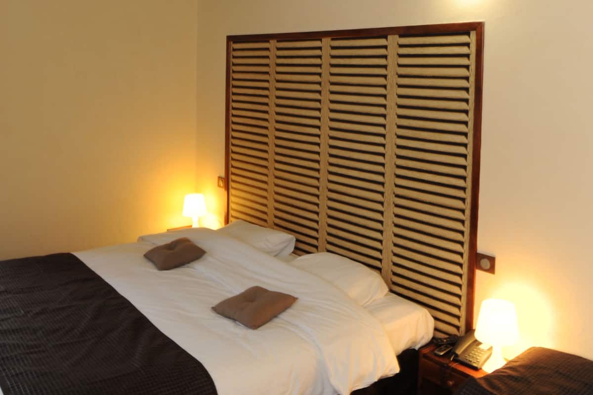 A hotel room with a wooden headboard, a bed with white linens, two bedside lamps, and a small bedside table with a lamp.