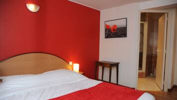 Une chambre d’hôtel comprenant un lit, une lampe fixée au mur, une petite table et une porte donnant accès à une salle de bain.