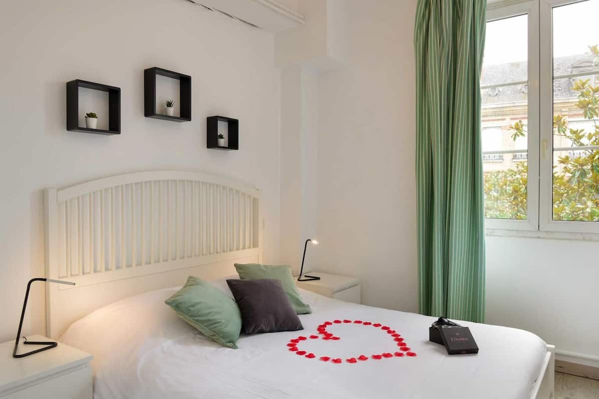 A neatly arranged bedroom with a bed, pillows, bedside lamps, and framed pictures on the wall.