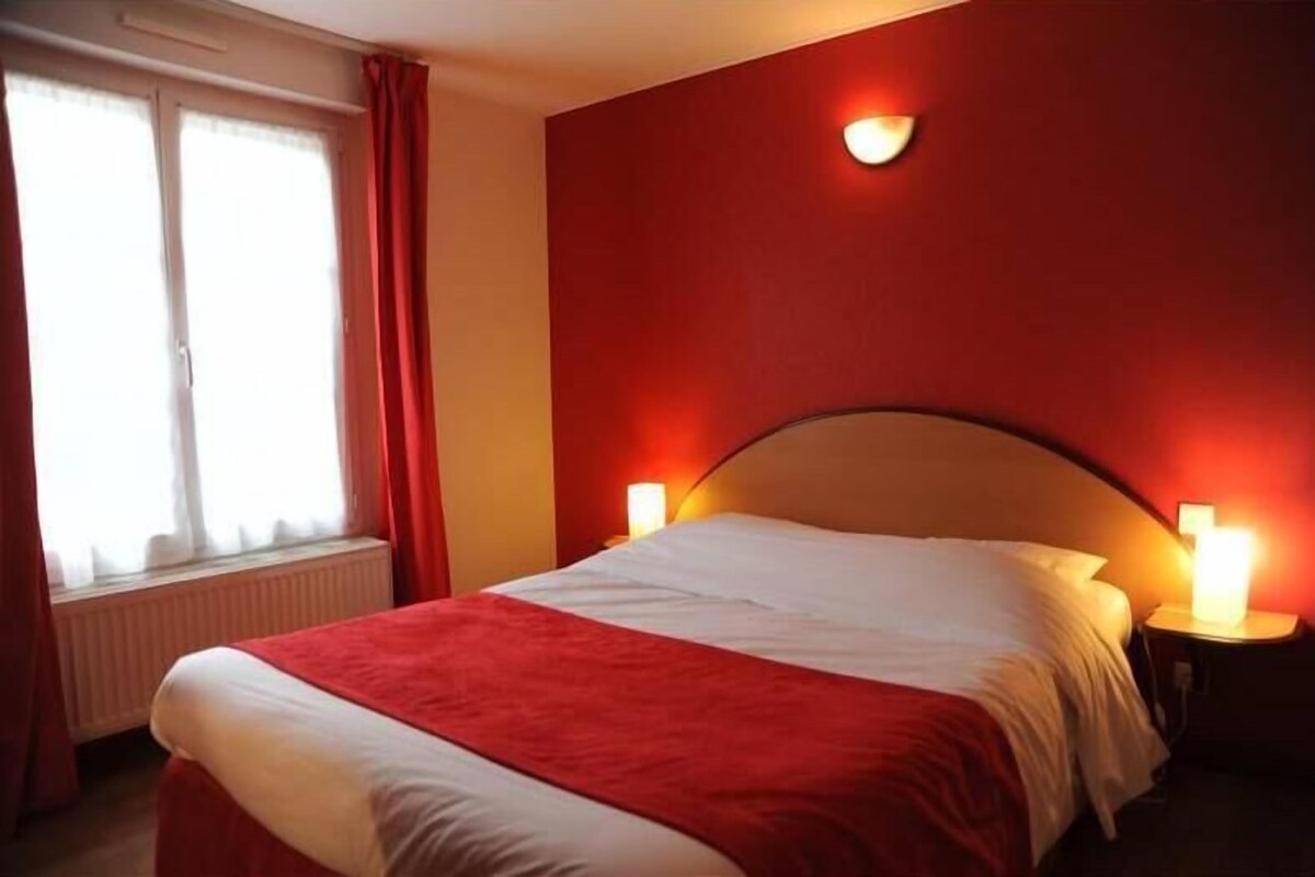 A hotel room with a bed, two bedside lamps, a window with curtains, and a wall-mounted light fixture.
