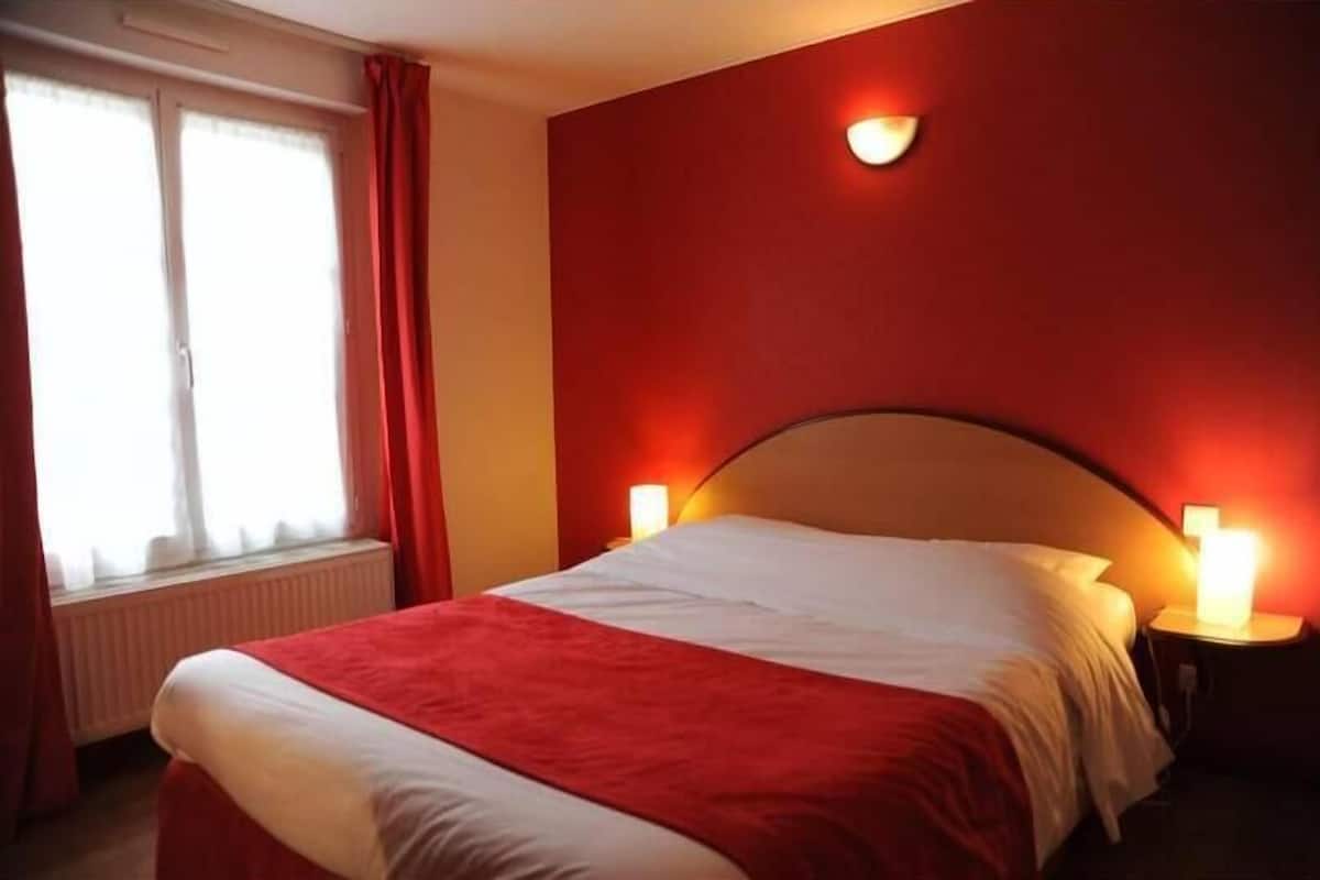 A hotel room with a bed, two bedside lamps, a window with curtains, and a wall-mounted light fixture.