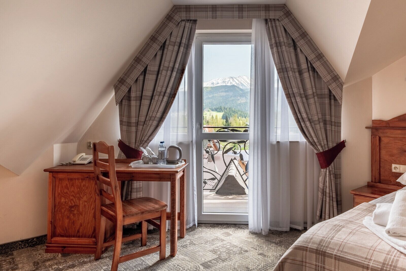 Un dormitorio con cama, un escritorio de madera con silla, teléfono y vistas a montañas a través de una ventana.