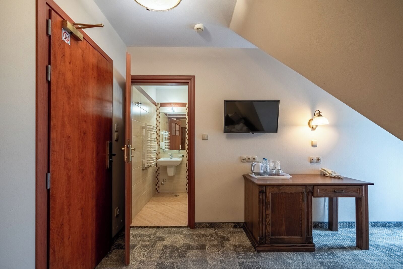 Una camera d'albergo con una porta in legno, una piccola scrivania con un telefono, una TV a schermo piatto montata a parete e un bagno visibile attraverso una porta aperta.