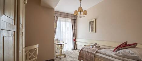 A bedroom with a bed, a chair, a small table, a window with curtains, and a chandelier.