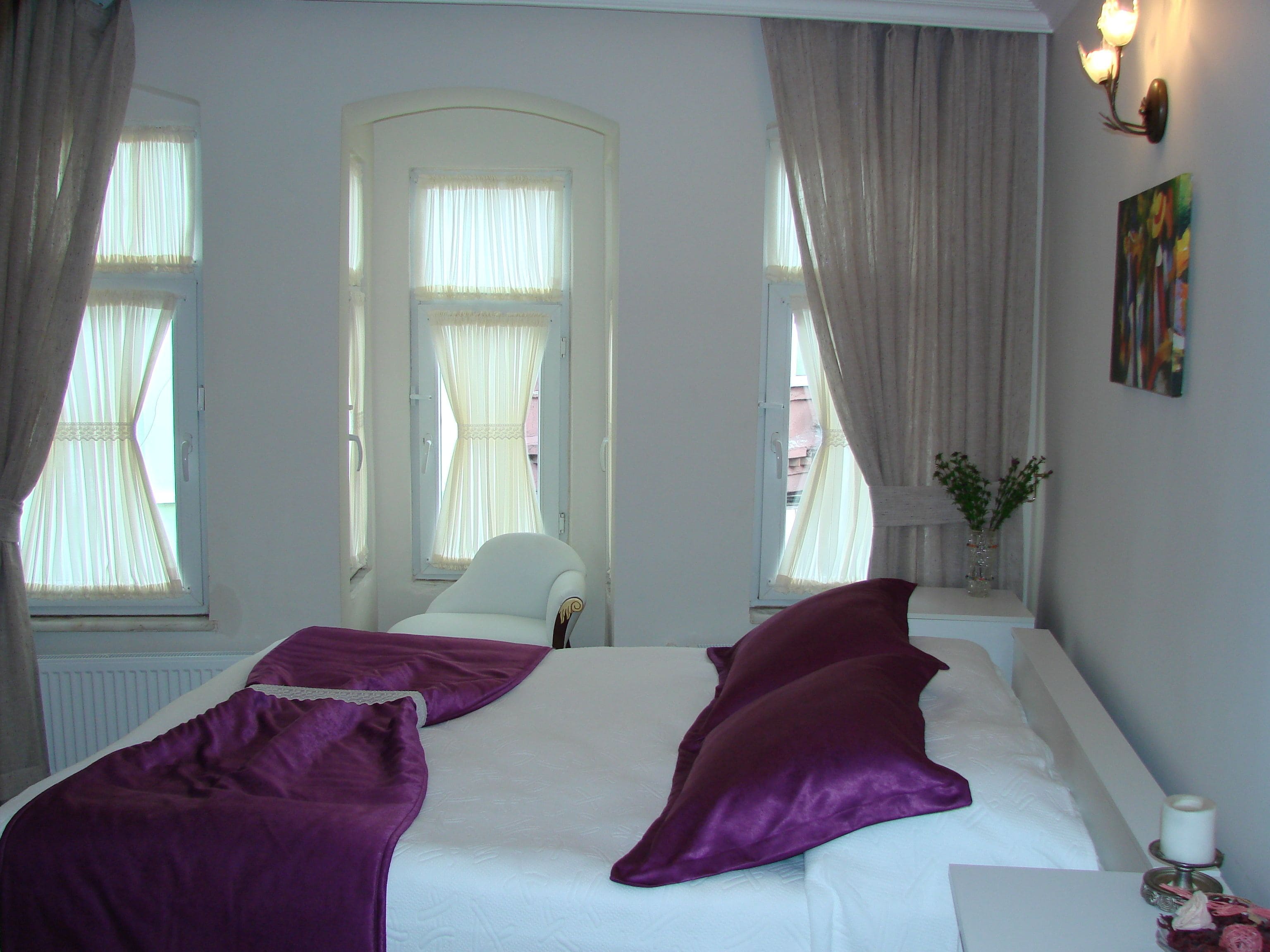 A bedroom with a bed, two windows with curtains, a chair, a bedside table with a vase, and a painting on the wall.