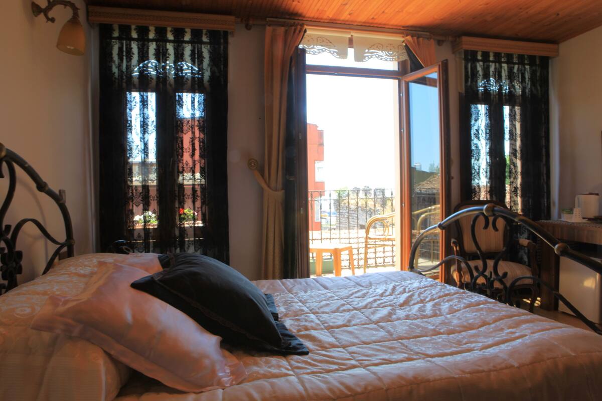 Un lit recouvert d’une courtepointe matelassée, un oreiller et une tête de lit en métal. Une fenêtre avec des rideaux est visible, et on aperçoit une chaise au fond.
